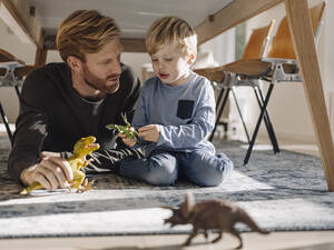 Father and son playing with dinosaur figures under the table at home - KNSF07024