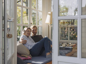 Portrait of smiling couple relaxing in sunroom at home - KNSF07035