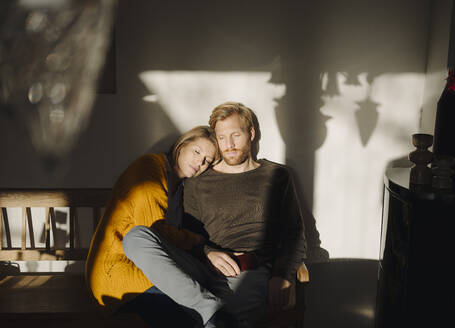 Relaxed couple sitting on bench in sunlight at home - KNSF07054