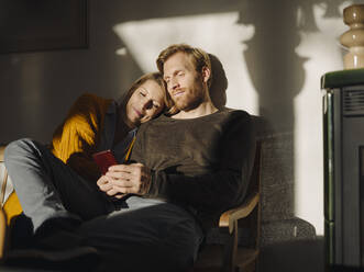 Relaxed couple sitting on bench in sunlight at home with man using cell phone - KNSF07056