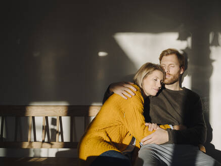 Relaxed couple sitting on bench in sunlight at home - KNSF07057