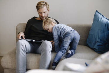 Vater und Sohn betrachten ein Buch auf der Couch zu Hause - KNSF07068