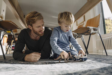 Father and son playing with dinosaur figure and toy car under the table at home - KNSF07120