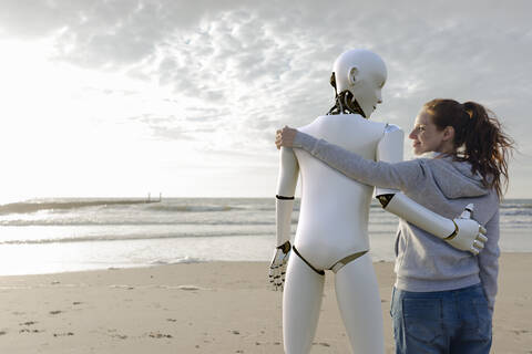 Smiling woman standing arm in arm with robot on the beach stock photo