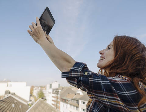 Smiling redheaded woman with tablet on rooftop terrace taking a selfie stock photo