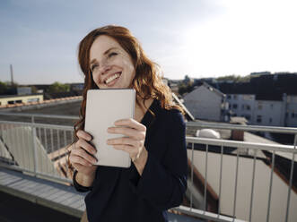 Glückliche rothaarige Frau mit Tablet auf einer Dachterrasse - KNSF07169