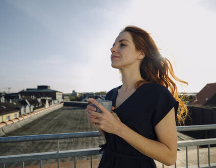 Smiling redheaded woman having a coffee break on rooftop terrace - KNSF07171