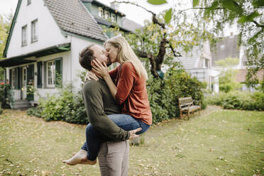 Happy couple kissing in garden, in front of their dream house - KNSF07221