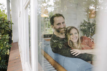 Happy couple sitting on couch, with arns around, smiling - KNSF07233