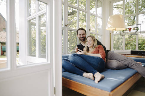 Glückliches Paar, das sich auf der Couch entspannt und ein Smartphone benutzt - KNSF07235
