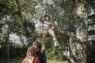 Son sitting on tree in garden with parents watching him - KNSF07241