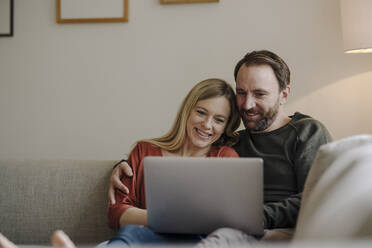 Couple sitting at home on couch, using laptop - KNSF07285