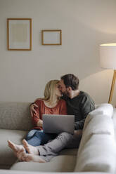 Couple sitting at home on couch, kissing, using laptop - KNSF07286