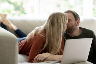 Couple sitting at home on couch, kissing, using laptop - KNSF07288