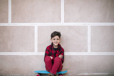 Porträt eines lächelnden Jungen auf einem Skateboard sitzend an einer Wand - GRCF00112