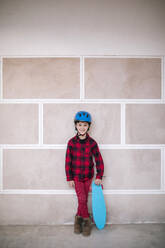 Porträt eines lächelnden Jungen mit Helm, der ein Skateboard an einer Wand hält - GRCF00114