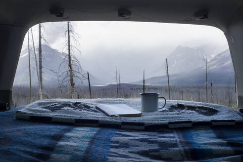 Tasse und Buch auf einer Decke im Kofferraum eines Autos mit dem Glacier National Park im Hintergrund - CAVF73564