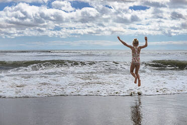 Rückansicht eines verspielten Mädchens, das in den Wellen von Virginia Beach gegen den bewölkten Himmel springt - CAVF73571