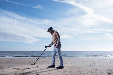 Side view of man using recreational metal detector at Horsey Beach against sky - CAVF73584