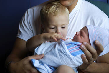 Kleinkind berührt neugeborenen Bruder, der in eine Decke eingewickelt und vom Vater getragen wird - CAVF73652