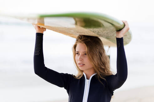 Woman looking away while carrying surfboard on head at beach - CAVF73656