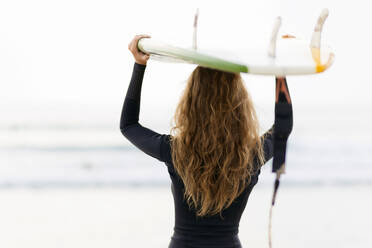 Rückansicht einer Frau mit Surfbrett auf dem Kopf am Strand - CAVF73657