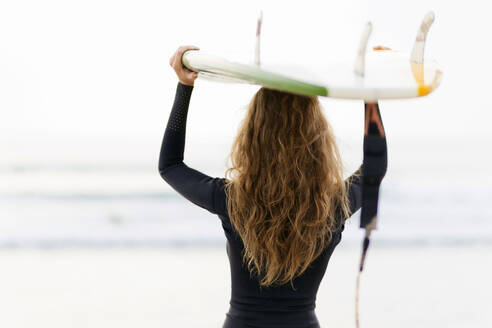 Rear view of woman carrying surfboard on head at beach - CAVF73657