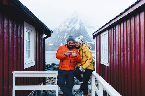 Tourist couple using smartphone on a porch at Hamnoy, Lofoten, Norway - DGOF00120