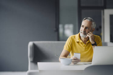 Porträt eines reifen Mannes, der mit Laptop und Smartphone am Tisch sitzt und in die Ferne schaut - KNSF07352