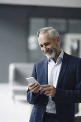 Portrait of smiling mature businessman in his office looking at smartphone - KNSF07366