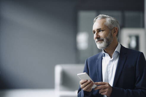 Portrait of smiling mature businessman with smartphone looking at distance - KNSF07367