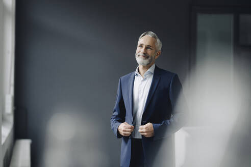 Portrait of smiling mature businessman wearing blue suit in office looking at distance - KNSF07370