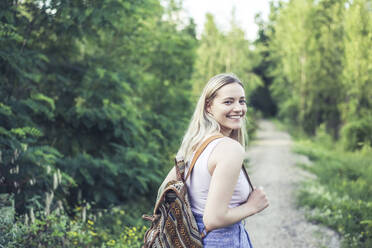 Porträt einer lächelnden jungen Frau mit Rucksack auf einem Waldweg - BFRF02182
