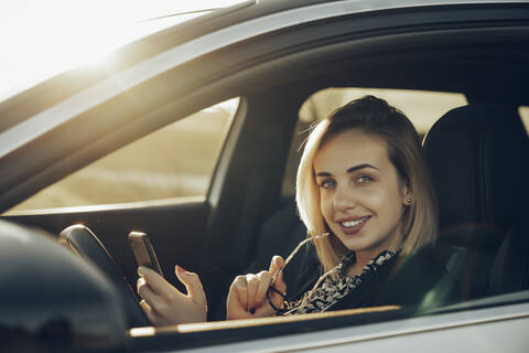 Porträt einer lächelnden jungen Frau mit Smartphone, die abends in ihrem Auto sitzt, lizenzfreies Stockfoto