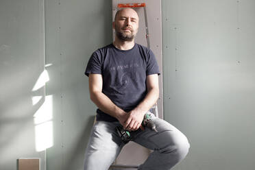 Porträt eines selbstbewussten Mannes mit einer tragbaren Bohrmaschine auf einer Baustelle zu Hause - KMKF01221