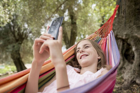 Mädchen in Hängematte in einem Olivenhain liegend, mit Smartphone - BFRF02192