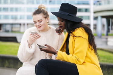 Freundinnen sitzen auf einer Bank in der Stadt und benutzen ein Mobiltelefon - DGOF00158