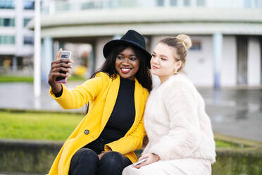 Glückliche Freundinnen sitzen auf einer Bank in der Stadt und machen ein Selfie - DGOF00161