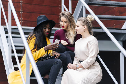 Freundinnen sitzen auf einer Treppe im Freien und schauen auf ihr Handy - DGOF00173