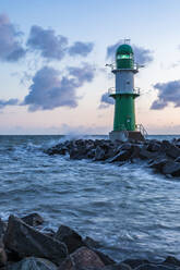 Germany, Mecklenburg-West Pomerania, Warnemunde, Lighthouse and sea waves crashing against rocks at dusk - WDF05723