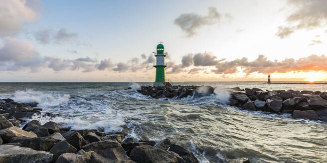 Deutschland, Mecklenburg-Vorpommern, Warnemünde, Leuchtturm und Meereswellen, die bei Sonnenuntergang gegen Felsen schlagen - WDF05724