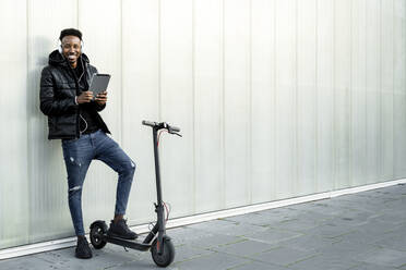 Portrait of happy man with headphones, tablet and e-scooter outdoors - RCPF00220