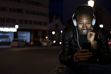 Mann in der Stadt bei Nacht mit Smartphone und Kopfhörern - RCPF00228