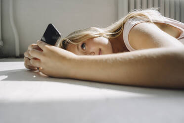Blonde junge Frau liegt auf dem Boden und benutzt ein Mobiltelefon - KNSF07387