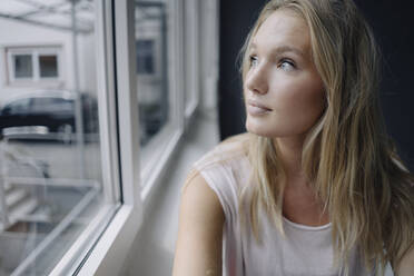 Porträt einer blonden jungen Frau, die aus dem Fenster schaut - KNSF07398
