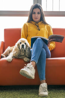 Portrait of teenage girl sitting with dog and book on the couch - DLTSF00429