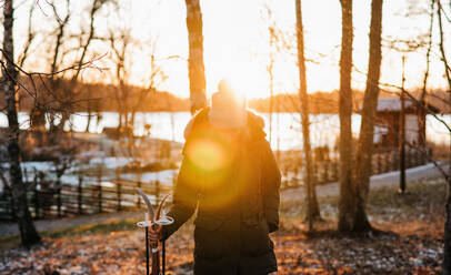 Woman walking through a snowy forest with skis at sunset - CAVF73733