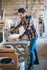 Carpenter sawing a board with a table saw in his workshop. - CAVF73899