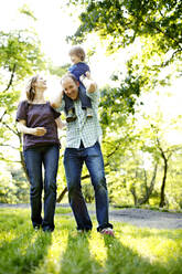 Dad holding son on shoulders in park with mom looking up - CAVF74048