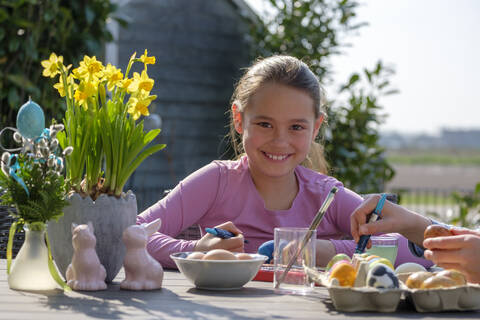 Porträt eines glücklichen Mädchens, das am Gartentisch sitzt und Ostereier bemalt, lizenzfreies Stockfoto
